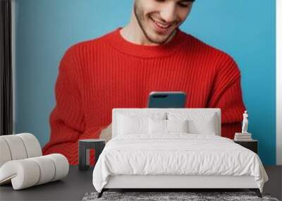 A young man smiling while using his smartphone to text or interact with social media. He is dressed in casual wear and has a cheerful demeanor Wall mural