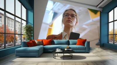 A young professional woman in a modern office environment, looking at her smartphone and engaging with digital technology. Wall mural