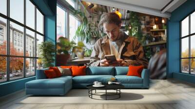 Man in casual attire sitting alone at a table, engrossed in his smartphone. Modern interior with indoor plants. Wall mural