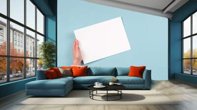 Female hands holding blank white paper sheet on light blue background. Top view. Mockup paper with copy space for text Wall mural