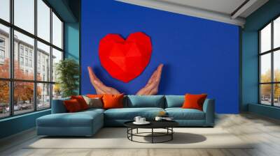 Female hands holding red polygonal heart on blue background. Top view Wall mural