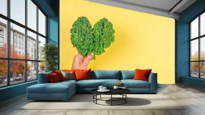 Woman hand holding green heart made of fresh curly kale cabbage leaves over yellow background. Love of vegetarian, vegan food, healthy organic food concept Wall mural