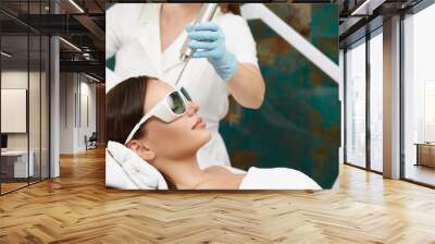 pretty girl lying in beauty salon and receiving laser theraphy for forehead wearing protection glasses Wall mural