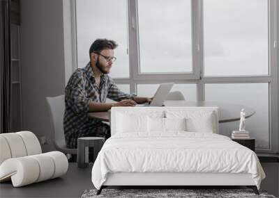 bearded man in glasses sits at home at a table with a laptop and works, self-isolation concept of online work on the Internet. Wall mural