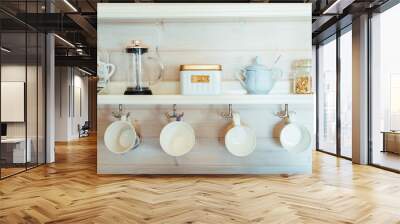 Kitchen wooden shelf with tea leaves in gold box and accessories, blue sugar bowl with spoon, strainer, press. Many colorful cups, mugs are hanging from hooks. Cozy interior in a country house Wall mural