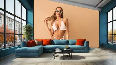 A tanned young woman in a white swimsuit and white sunglasses laughs on a beige background. Wall mural