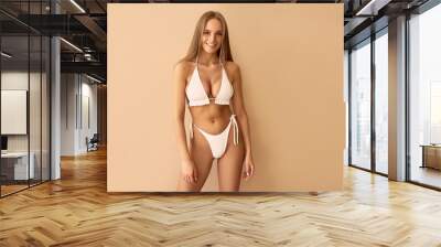 A young beautiful tanned woman in a white swimsuit stands near a beige background. Wall mural