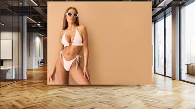 Beautiful young woman in a white swimsuit and white sunglasses on a beige background. Wall mural