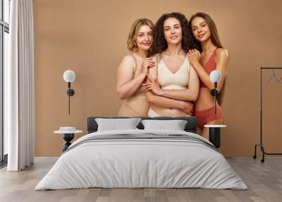 Feminine beauty. Group of three beautiful caucasian women with different body types and skin color standing in embrace in studio. Young ladies wearing lingerie posing over beige background. Wall mural