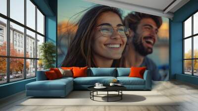 beautiful young woman with long brown hair and wearing eyeglasses is smiling and looking off into the distance. A handsome man with a well-groomed beard is also in the photo, laughing Wall mural