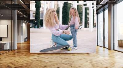 Mother and daughter sit on skateboard. Woman with unusual, lush and long hair. Small girl and woman smiles and happy. same colour  of clothes, get fun outdoor in weekend. Small kid study skating.  Wall mural