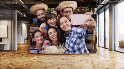 Brazilian June Party, typical celebration in Brazil. Happy men and women together in typical plaid shirt and straw hat taking a photograph with cell phone. Rustic wooden decor with flags. Wall mural