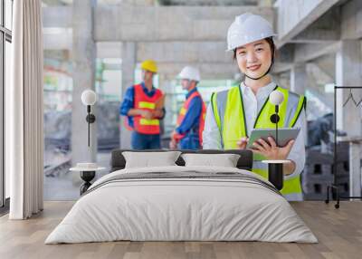 asian female engineer holds digital tablet,smiling and posing to camera,background the colleagues team inspecting under construction building Wall mural