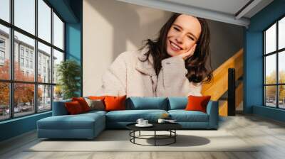 Portrait of smiling young woman in coat with brunette curly hair looking at camera indoors Wall mural