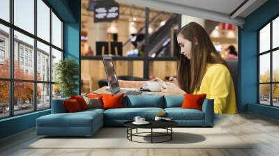 Young female freelancer in yellow jacket working in a cafe. Woman drinking coffee, browsing via moble phone and working with computer. Freelance work Wall mural