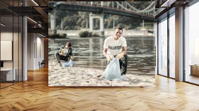 Cleaning up trash. Two smiling responsible students feeling cheerful while cleaning up trash left behind on the beach Wall mural