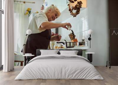 Joyful senior woman in apron cooking dinner in kitchen Wall mural