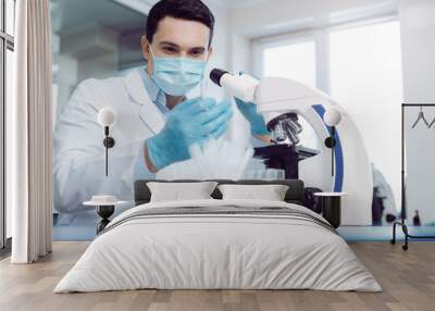 Modern research. Professional smart male scientist wearing uniform and looking at the test tube while working in the laboratory Wall mural