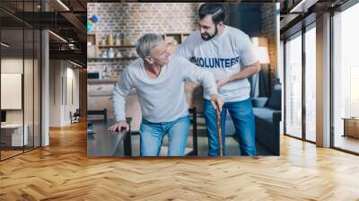 So helpful. Handsome joyful dark-eyed man smiling and helping an old man while he standing up Wall mural