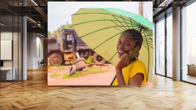 young beautiful african lady using an umbrella to protect herself under a very sunny weather and making calls with her mobile phone Wall mural