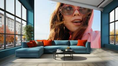 Stylish woman in sunglasses and pink hoodie poses confidently on the beach, making eye contact with the camera Wall mural