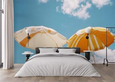 Two beach umbrellas in vibrant orange and white colors against a bright blue sky with scattered clouds Perfect for summer themes and vacation imagery Wall mural