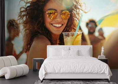 A young brunette woman wearing a hat, sunglasses and a swimsuit on the beach, smiling and holds a refreshing juicy cocktail in her hand. Day sunlight reflection. Beach party and summer leisure concept Wall mural