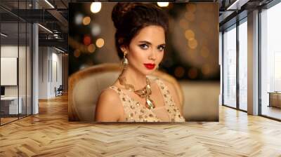 Luxurious portrait of elegant woman with wedding hairstyle and makeup. Beautiful brunette girl with golden jewelry in prom dress sitting on modern chair over bokeh lights xmas decorations. Wall mural