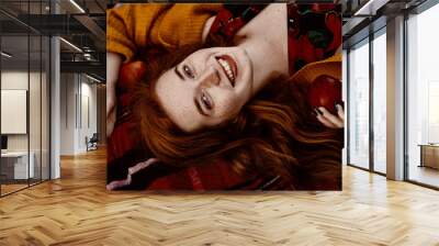 Close up flat lay, top view autumn portrait of young beautiful fashionable   happy smiling natural redhead woman with freckles, long curly hair, holding red apple, laying on plaid. Close up portrait Wall mural