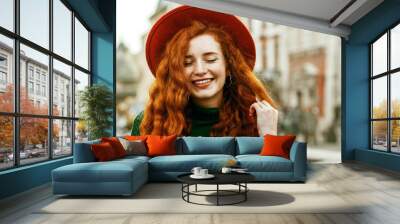 Close up portrait of young beautiful fashionable happy smiling redhead woman with freckles, very long curly hair, wearing green turtleneck, orange hat, golden wrist watch, posing in street. Copy space Wall mural