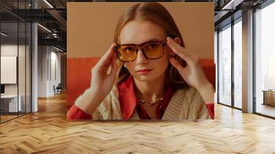 Fashionable confident woman wearing trendy yellow glasses with leopard frames, sitting in armchair, posing on beige background. Close up portrait. Copy, empty space for text Wall mural