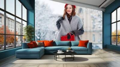 Happy smiling woman enjoying winter holidays, walking in snow covered street of European city. Model wearing beret, grey scarf, coat, gloves, with red quilted bag. Copy, empty space for text  Wall mural