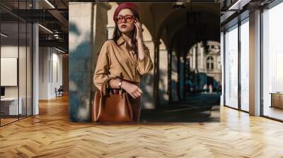 Outdoor, street style, fashion portrait of elegant, luxury woman wearing faux leather dark red beret, sunglasses, beige shirt, wrist watch, holding brown handbag, posing in European city. Copy space Wall mural