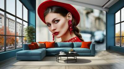 Outdoor close up portrait of young beautiful fashionable confident woman with red lips makeup, wearing total red look: hat, floral sequin earrings, dress. Model posing in street. Copy space for text Wall mural