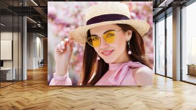 Outdoor close up portrait of young beautiful happy smiling girl wearing trendy yellow color sunglasses, straw boater hat, earrings, off shoulder pink blouse. Spring, summer fashion concept Wall mural