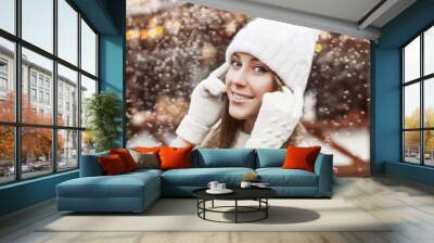 Outdoor close up portrait of young beautiful happy smiling woman wearing knitted white winter beanie hat, gloves, sweater, posing in festive Christmas fair in european city. Winter holidays concept Wall mural