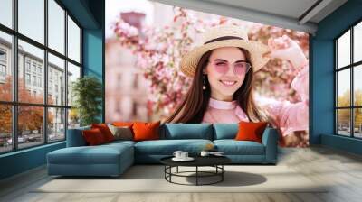 Outdoor close up portrait of young beautiful happy smiling woman wearing stylish pink cat eye sunglasses, straw hat, earrings, blouse, posing in street of city. Copy, empty space for text Wall mural