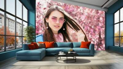 Outdoor close up portrait of young beautiful smiling girl wearing stylish pink color sunglasses, earrings, blue jacket, posing in street near blooming tree. Spring fashion concept. Copy, empty space Wall mural