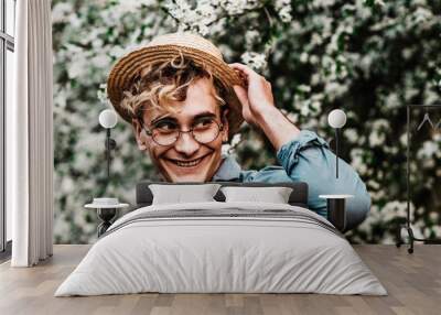 Outdoor close up portrait of young handsome happy smiling man wearing glasses and straw hat posing in the blooming garden Wall mural