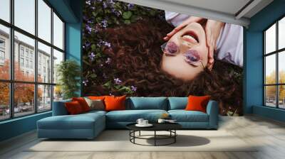 Outdoor flat lay, top view close up portrait of young beautiful happy smiling lady with long curly hair, wearing stylish purple sunglasses, lying on the meadow with violets. Copy, empty space  Wall mural