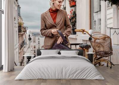 Outdoor full-length autumn portrait of young happy smiling fashionable lady wearing trendy leather beret, beige checkered blazer, wide trousers, white sneakers, holding bag, posing in Parisian street Wall mural