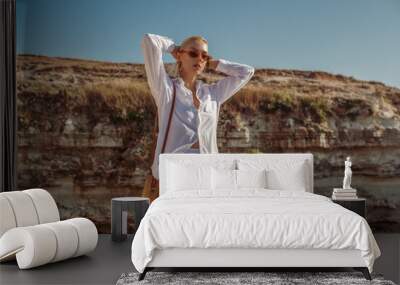 Outdoor summer fashion portrait of elegant woman wearing white linen suit, sunglasses, with round wicker bag, posing on rocks near sea. Copy, empty space for text Wall mural