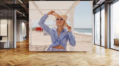 Outdoor summer fashion portrait of happy smiling woman wearing stylish straw hat, sunglasses, chain necklace, blue linen shirt posing on sea beach. Copy, empty space for text Wall mural