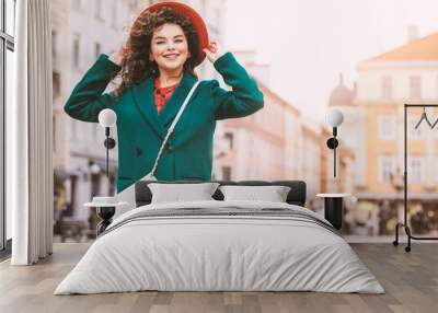 Young beautiful happy smiling lady wearing orange hat, green coat, with white cross body bag posing in street of European city. Copy, empty space for text Wall mural