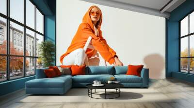 Young confident blonde girl wearing trendy orange hoodie, color sunglasses, posing on white background. Studio fashion portrait. Copy, empty space for text Wall mural