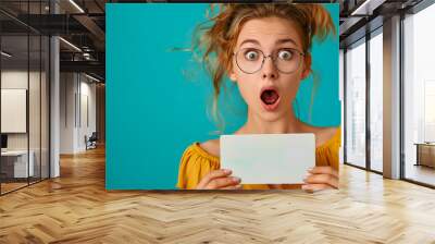 Woman with blonde hair and glasses holding blank card in front of blue background Wall mural