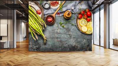 Tasty asparagus cooking with tomatoes, lemon and seasoning, preparation on rustic kitchen table with wooden spoon, top view, border. Dark style Wall mural