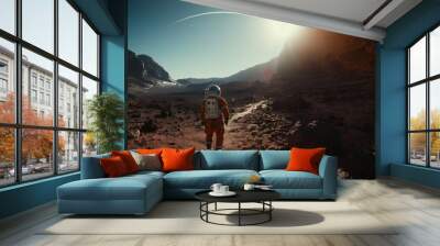 An astronaut in a vibrant orange suit hikes across a rocky Martian terrain, the horizon aglow with the light of a distant sun. Wall mural