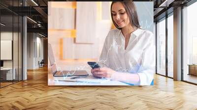 Attractive businesswoman in white shirt using smart phone to optimize trading strategy at corporate finance fund. Forex chart hologram over modern office background Wall mural