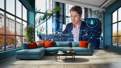 Handsome businessman in suit at workplace working with laptop to hire new employees for international business consulting. HR, social media hologram icons over office background Wall mural